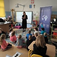 dzieci siedzą podłodze w sali lekcyjne. Kobieta stoi przed dziećmi i prowadzi zajęcia