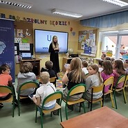 dzieci siedzą podłodze w sali lekcyjne. Kobieta stoi przed dziećmi i prowadzi zajęcia