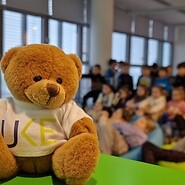 pluszowy misiek na pierwszym planie, w tle dzieci siedzą na podłodze