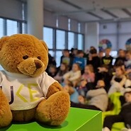 pluszowy misiek na pierwszym planie, w tle dzieci siedzą na podłodze